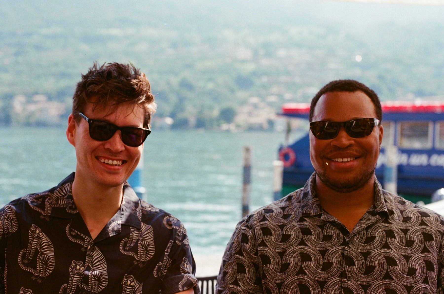 Two men wearing sunglasses with a scenic background of water and greenery.