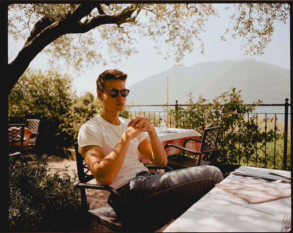 Man sitting outdoors under a tree, wearing sunglasses and a white shirt.