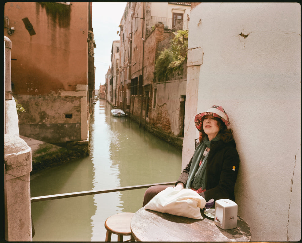 Person sitting by a canal in an urban setting with buildings and boats.